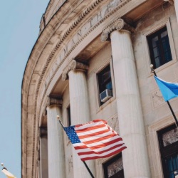 新古典主义政府大楼的特写，有大柱子和石头立面. Several flags, including the American flag, are visible in front of the building against a clear blue sky.